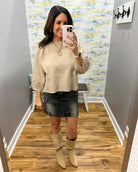 A woman takes a mirror selfie in a fitting room wearing a beige knit long-sleeve sweater, a dark gray denim mini skirt, and beige suede knee-high boots.