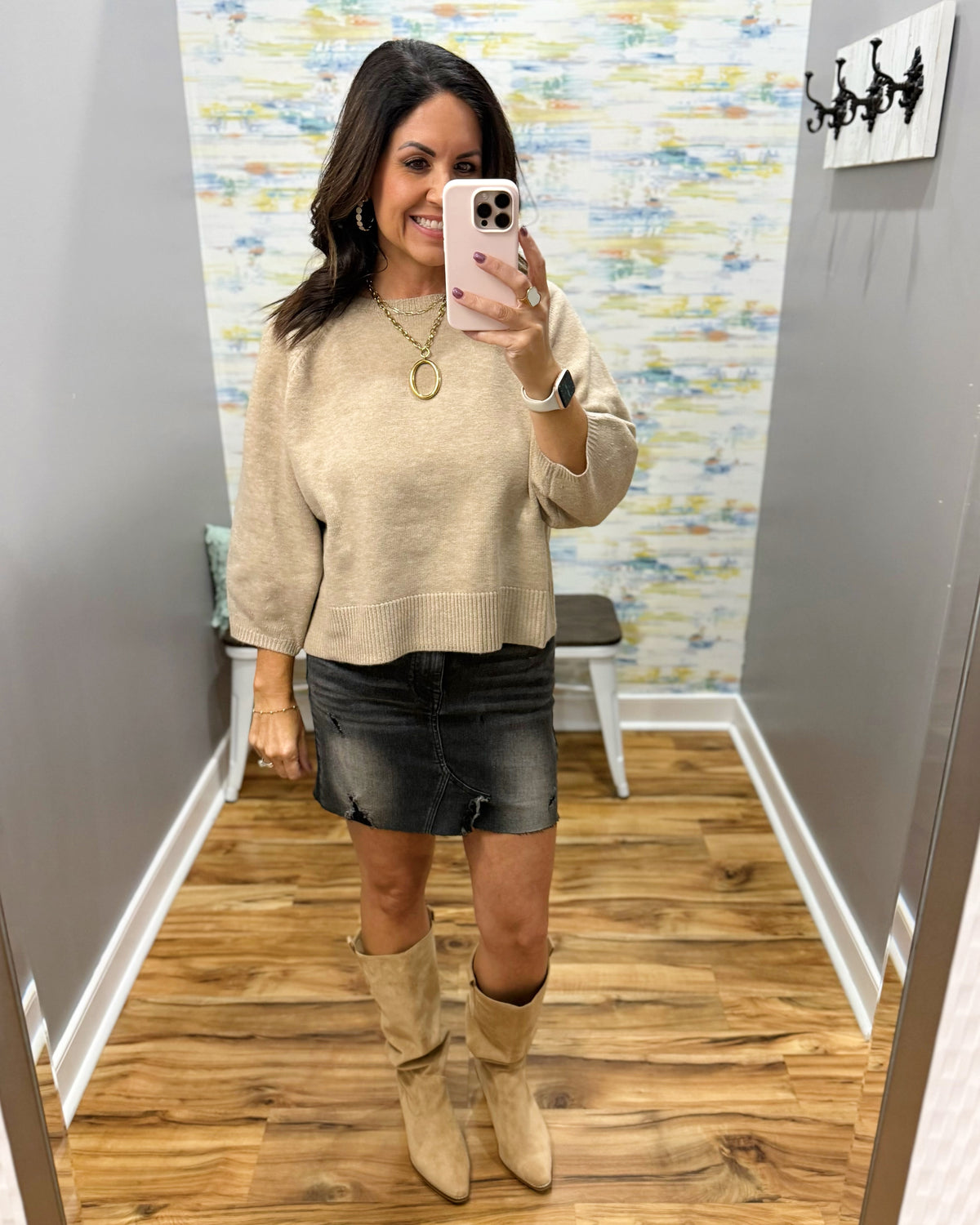 A woman takes a mirror selfie in a fitting room wearing a beige knit long-sleeve sweater, a dark gray denim mini skirt, and beige suede knee-high boots.