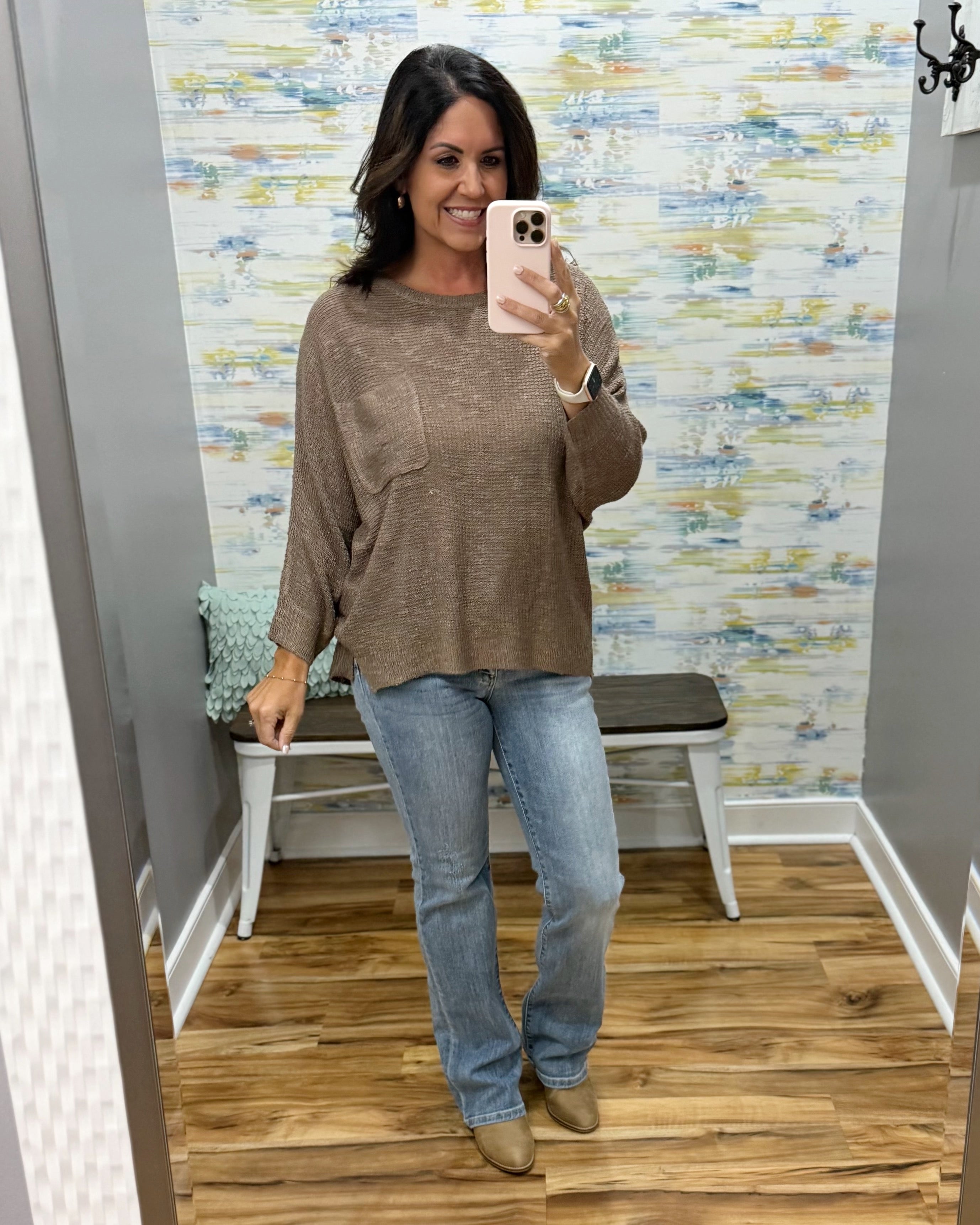 A woman takes a mirror selfie in a fitting room wearing a brown knit long-sleeve top, light blue jeans, and tan boots.