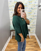 A woman takes a mirror selfie wearing a dark green, oversized three-quarter sleeve top and blue jeans in an indoor hallway with wood floors and a patterned wall.