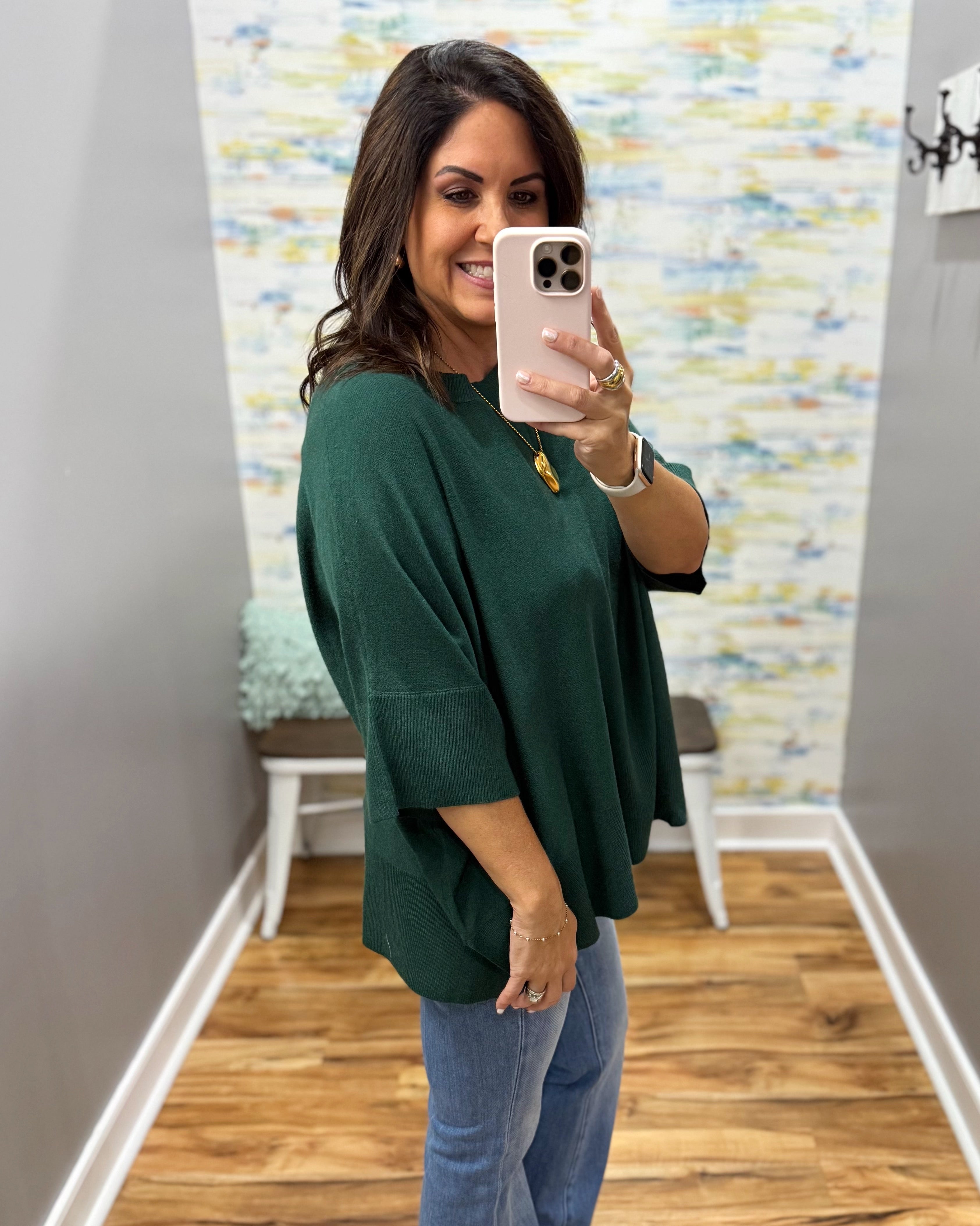 A woman takes a mirror selfie wearing a dark green, oversized three-quarter sleeve top and blue jeans in an indoor hallway with wood floors and a patterned wall.
