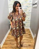 A woman takes a mirror selfie in a boutique showing a short-sleeve floral dress with puff sleeves, a V-neck, and brown Mary Jane shoes.