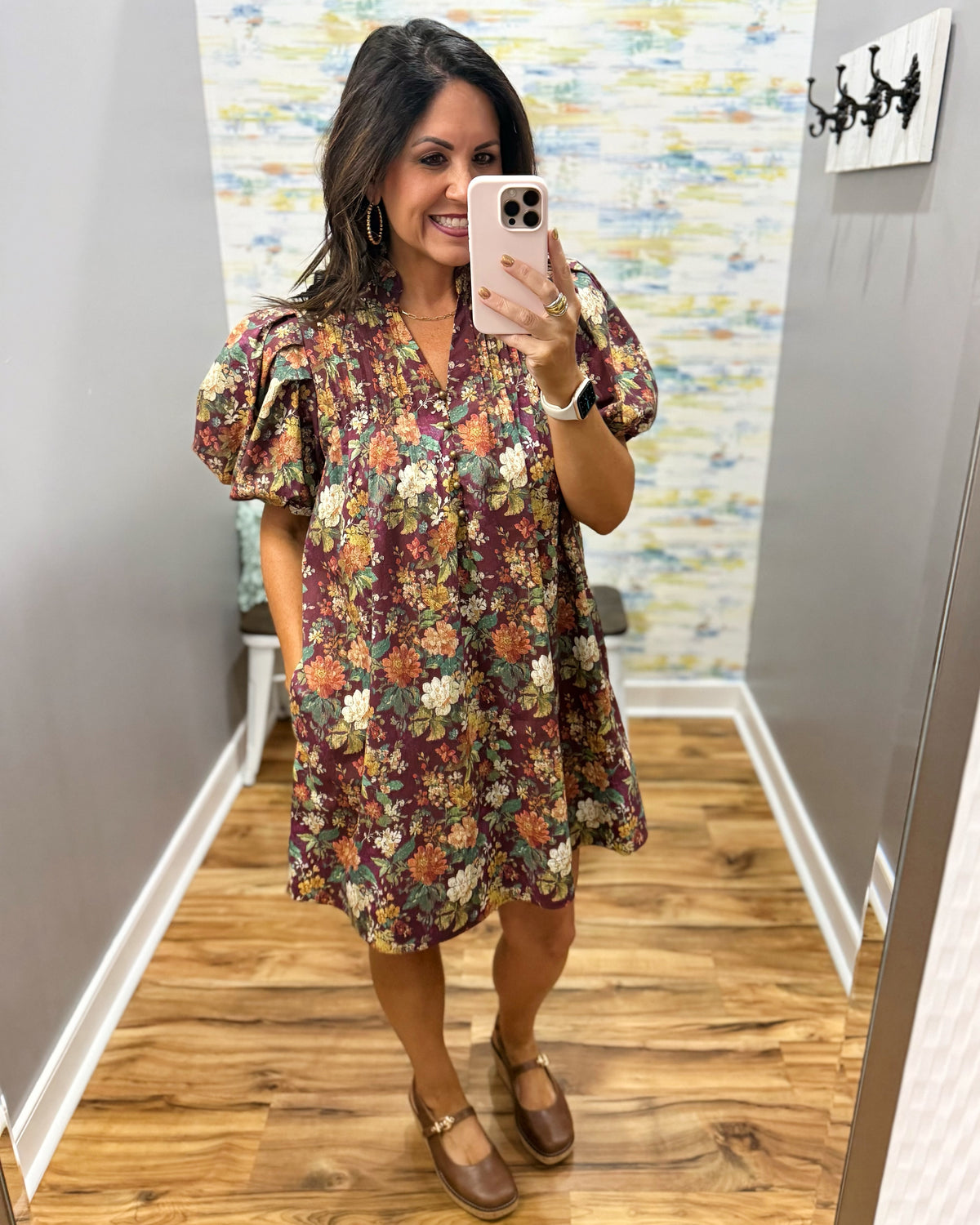 A woman takes a mirror selfie in a boutique showing a short-sleeve floral dress with puff sleeves, a V-neck, and brown Mary Jane shoes.