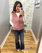 A woman takes a mirror selfie in a fitting room wearing a dusty pink sleeveless puffer vest over a white long-sleeve top, blue flare jeans, and beige shoes.