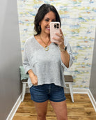 Woman taking a mirror selfie wearing a gray textured top and denim shorts in a room with patterned wallpaper.