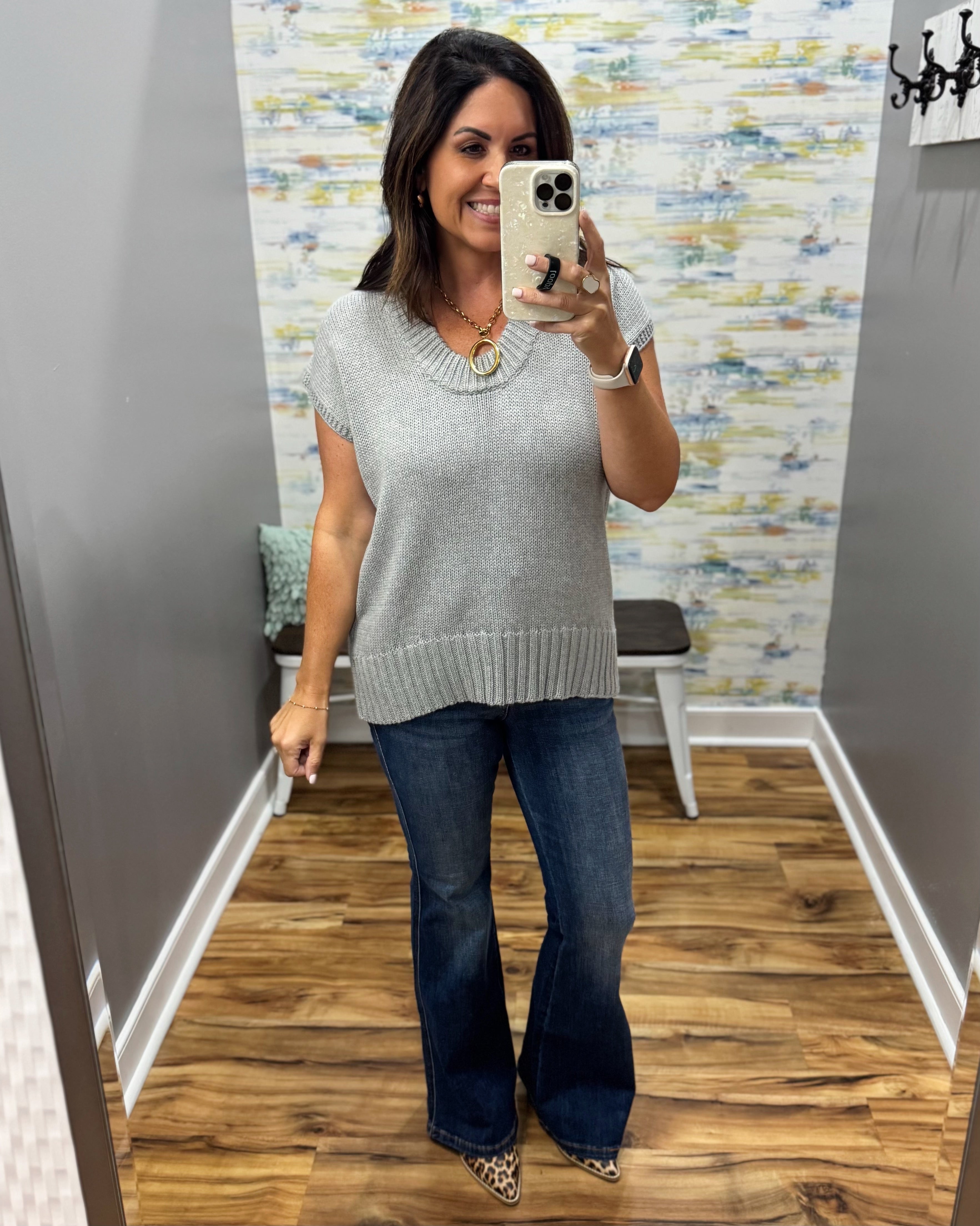 A woman takes a mirror selfie in a fitting room wearing a light gray ribbed knit sweater vest with short sleeves, paired with dark blue flare jeans and leopard print shoes.