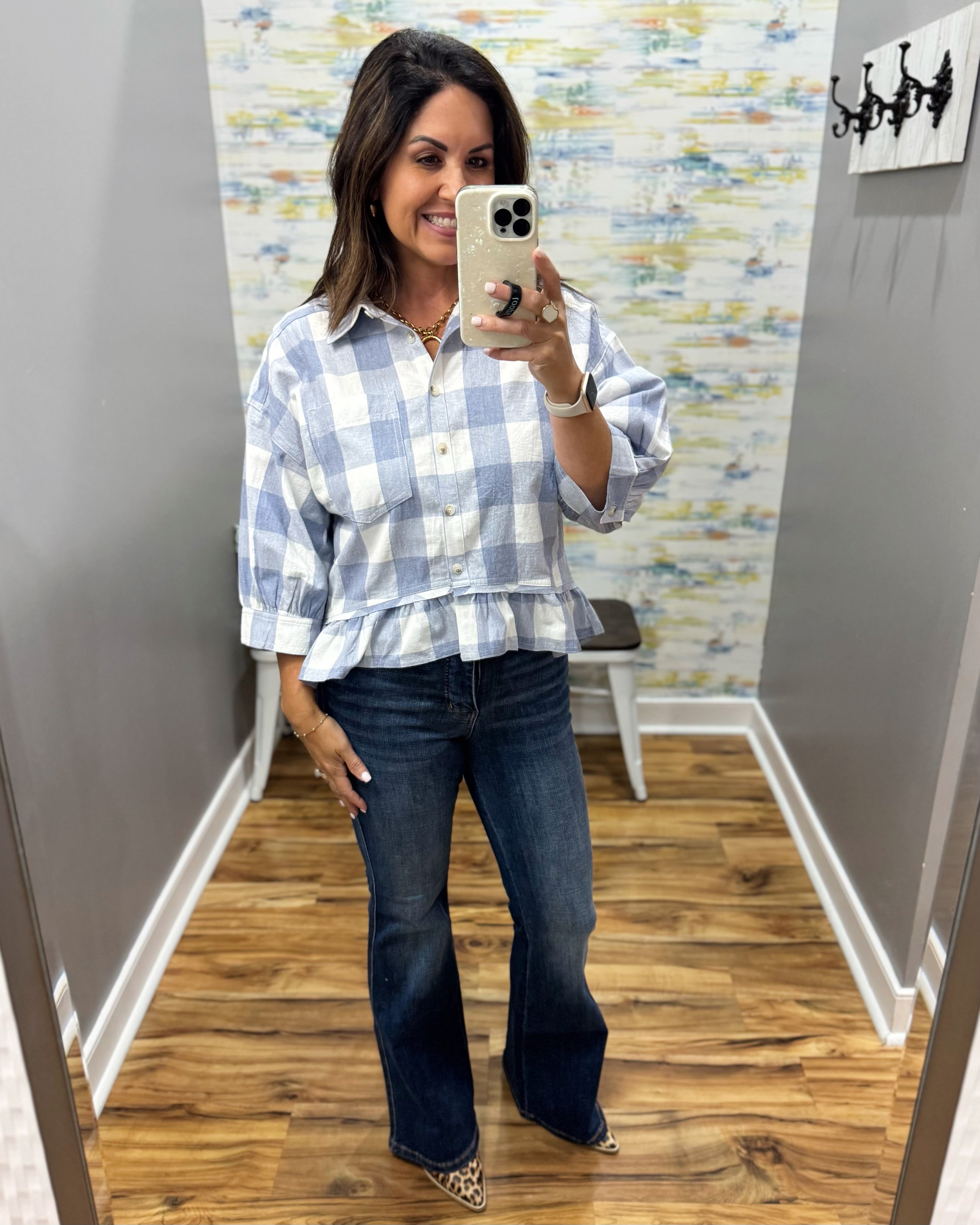 A woman takes a mirror selfie in a fitting room wearing a light blue and white checkered peplum blouse with dark blue flare jeans.