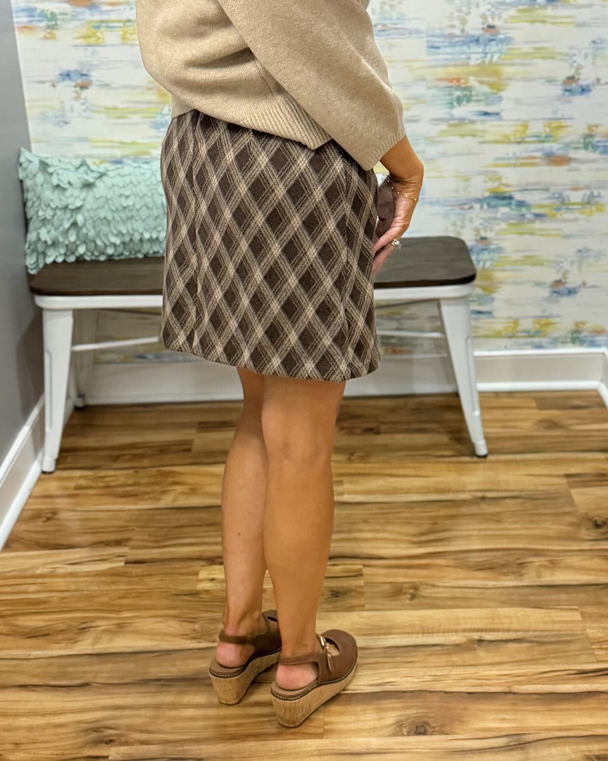 A person wearing a brown and beige plaid mini skirt, a beige sweater, and Corkys Pity Party in Cognac, standing in a room with a wood floor and a bench in the background.