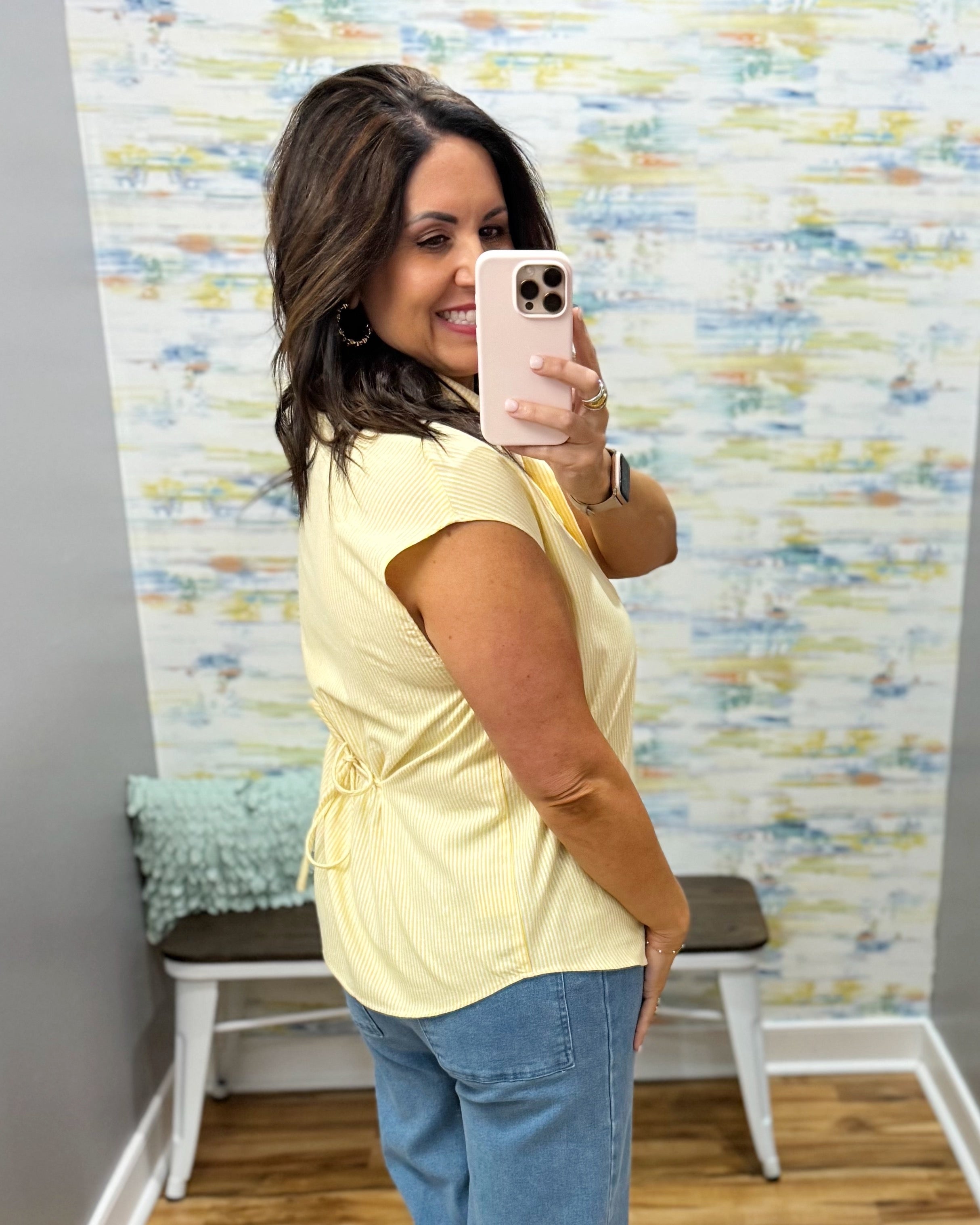 Woman taking a mirror selfie wearing a yellow top and blue jeans in a room with colorful wall paper.