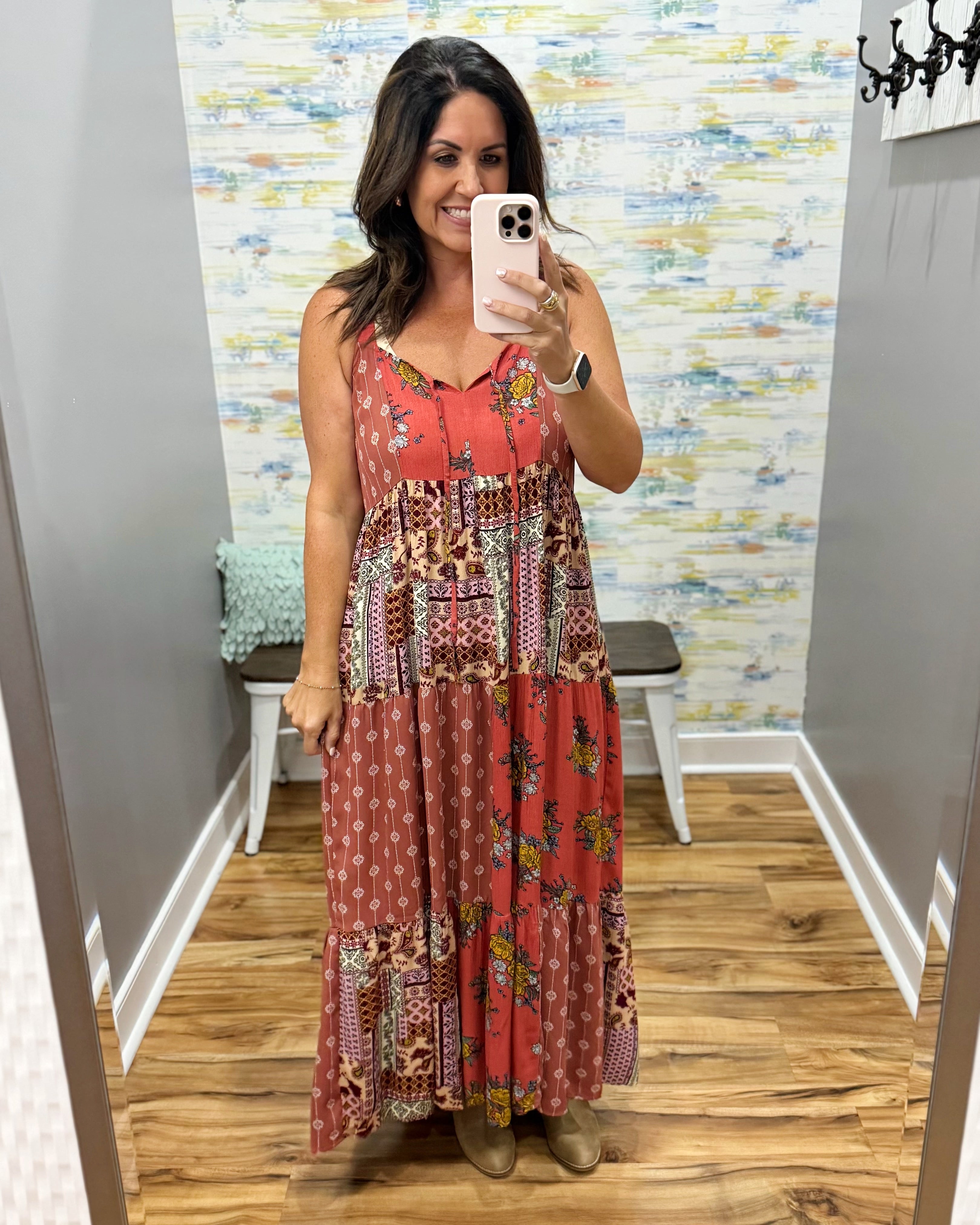 A woman takes a mirror selfie in a fitting room wearing a sleeveless patchwork maxi dress with a rust red base and multicolor floral and geometric prints. The background features a patterned wallpaper, wooden floors, and a small bench.