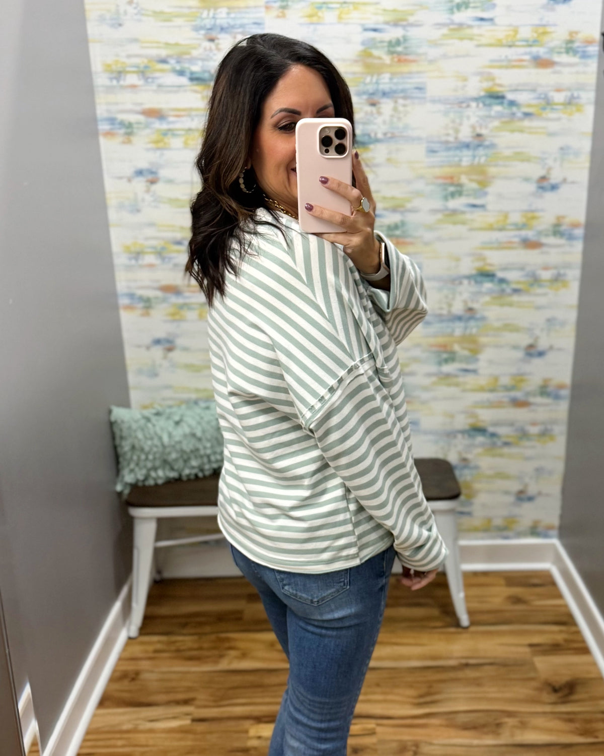 A woman takes a mirror selfie wearing a light green and white striped long-sleeve top, blue jeans, and a light pink phone case in a home setting with a patterned wallpaper background.