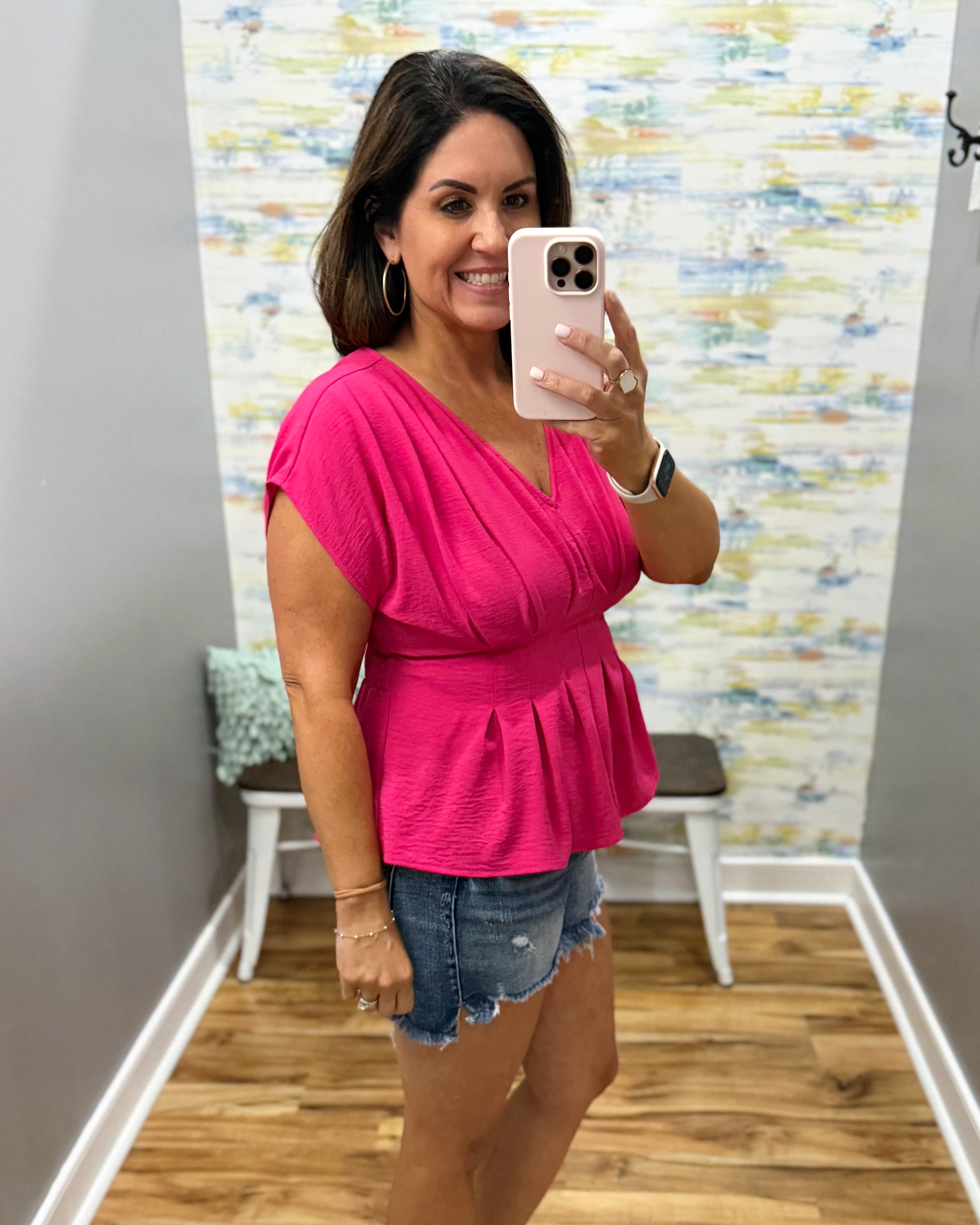 Woman taking a mirror selfie wearing a pink top and denim shorts in a room with a colorful wall.