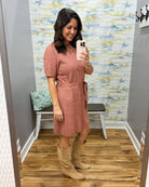 A woman takes a mirror selfie in a fitting room wearing a dusty rose short-sleeve knit dress with a tie waist and tan knee-high boots.