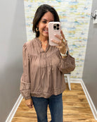 A woman takes a mirror selfie wearing a dusty rose long-sleeve blouse with a gathered, pleated neckline and blue jeans in a home setting with wood floors and gray walls.