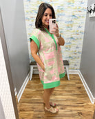 Woman taking a mirror selfie wearing a pink and green dress in a room with a patterned wall.