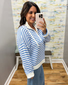 A woman takes a mirror selfie wearing a blue and white striped knit quarter-zip sweater with a white collar, paired with blue jeans in an indoor room with wood floors and a patterned wall.