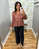 A woman takes a mirror selfie wearing a rust-colored floral long-sleeve peplum blouse and dark wide-leg pants in a home interior with wood floors and a patterned wall.