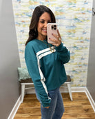 A woman takes a mirror selfie wearing a teal long-sleeve sweatshirt with white athletic stripes on the chest and sleeves, paired with blue jeans.