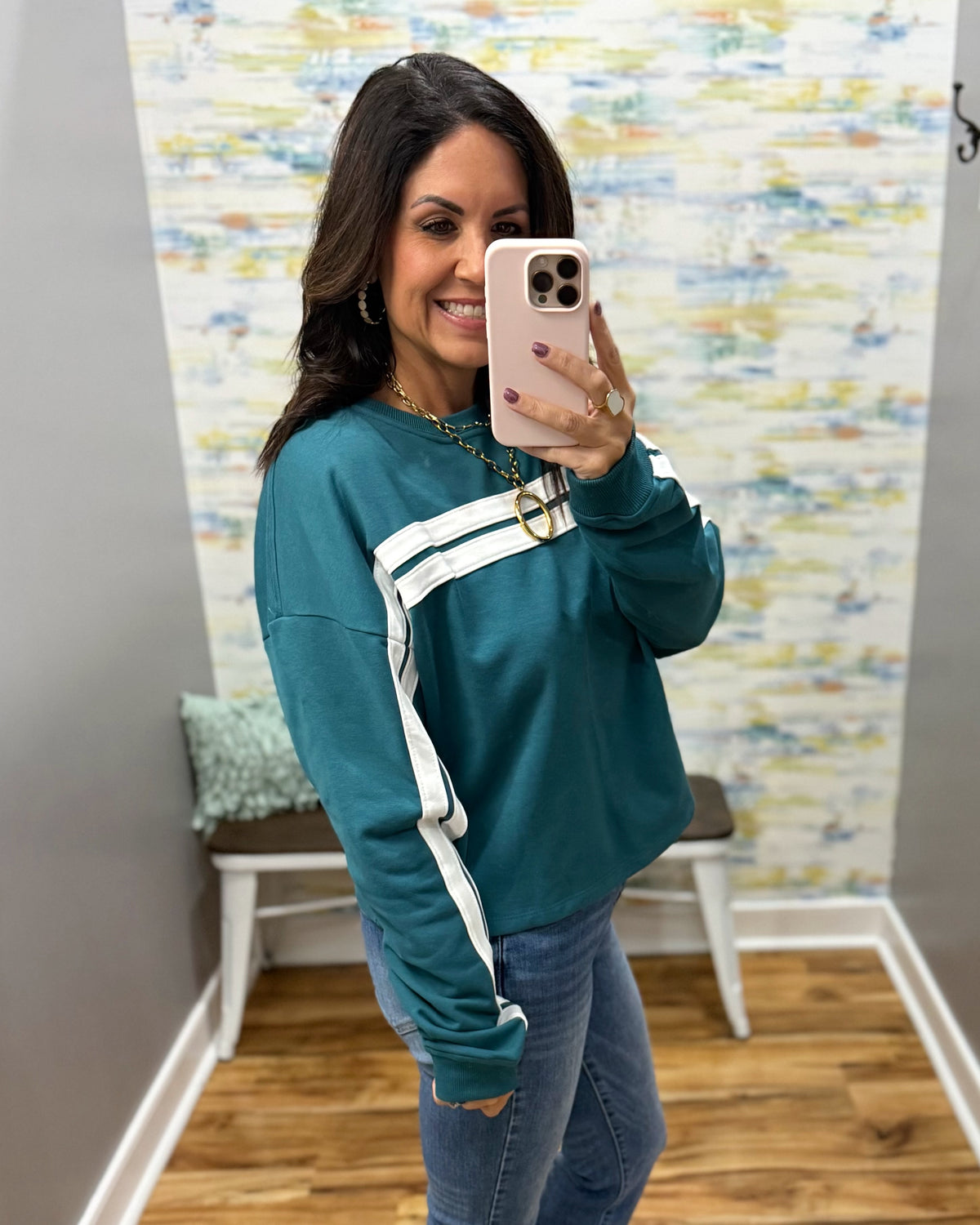 A woman takes a mirror selfie wearing a teal long-sleeve sweatshirt with white athletic stripes on the chest and sleeves, paired with blue jeans.
