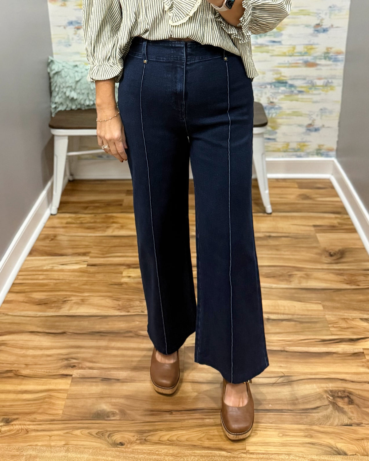 A person wearing dark navy wide-leg jeans with contrast light stitching, paired with a striped blouse and brown shoes, standing on a wood floor in an indoor room.