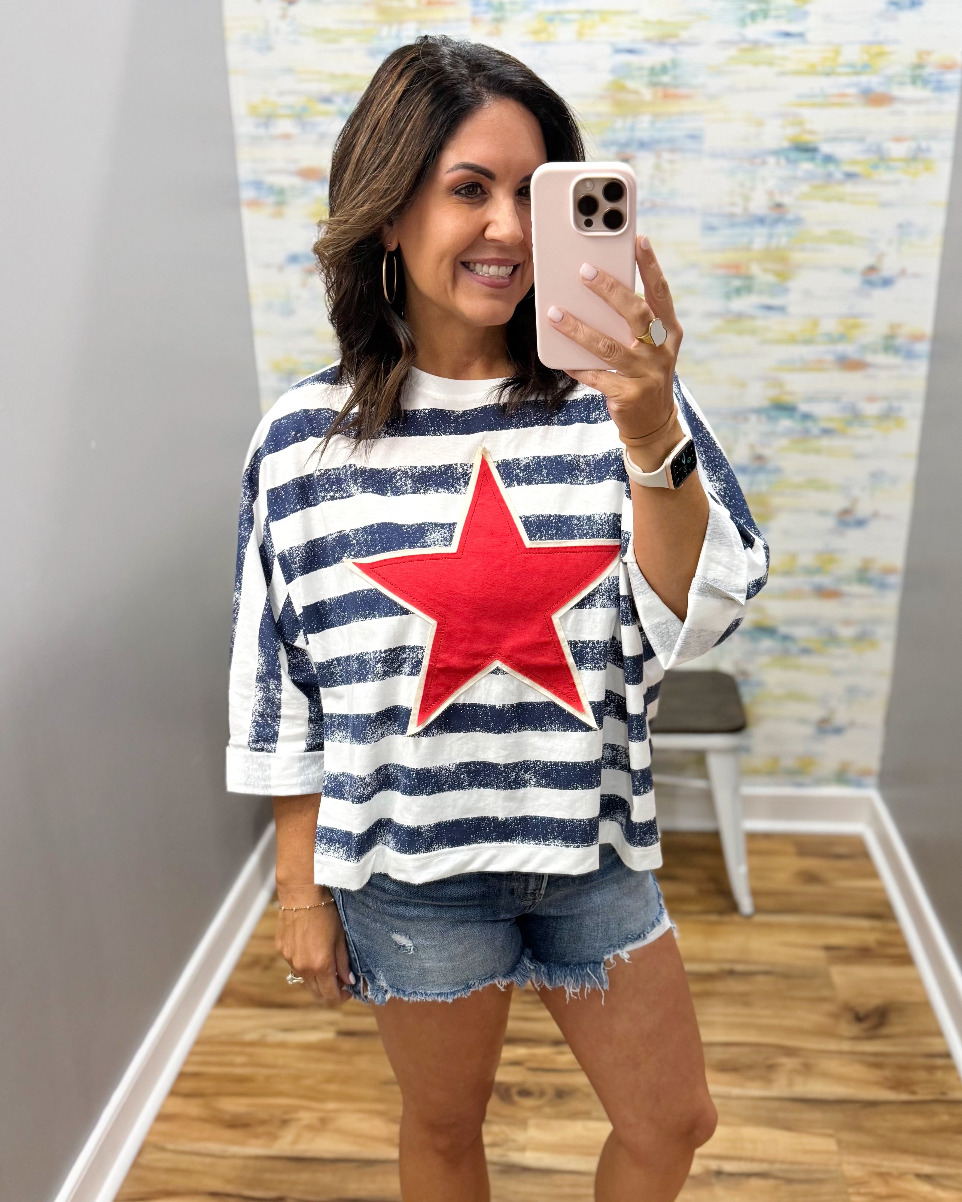 Woman wearing a striped shirt with a red star, taking a mirror selfie.