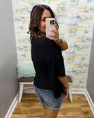Woman taking a mirror selfie wearing a black top and denim shorts in a room with a textured wall.