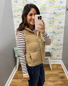 A woman takes a mirror selfie wearing a tan quilted puffer vest over a black and white striped long-sleeve shirt, dark blue jeans, and tan boots in a home fitting room.