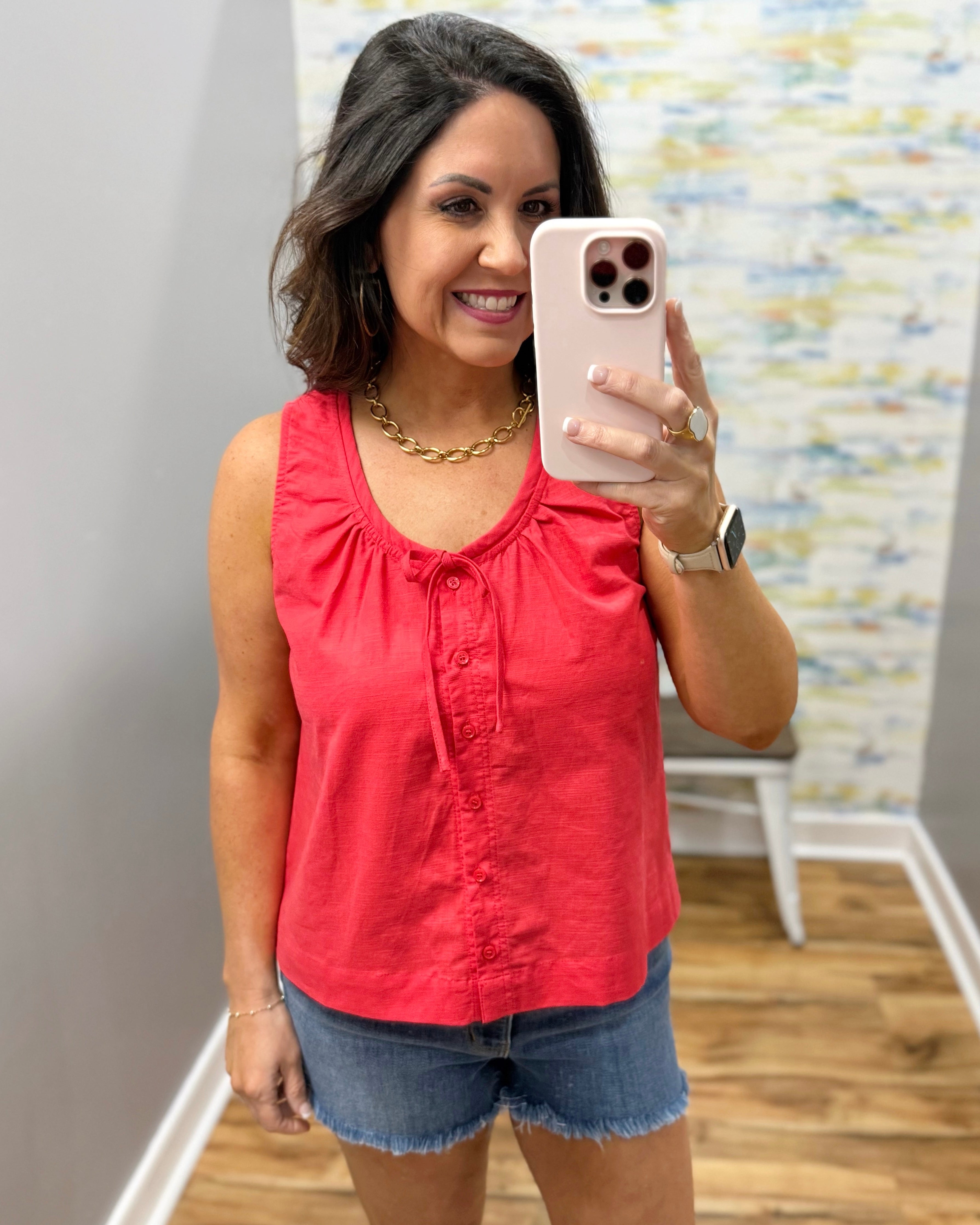 Woman taking a mirror selfie wearing a red sleeveless top and denim shorts.