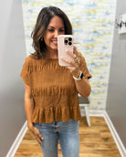 A woman takes a mirror selfie wearing a brown smocked short-sleeve top with a ruffled hem and light wash blue jeans.