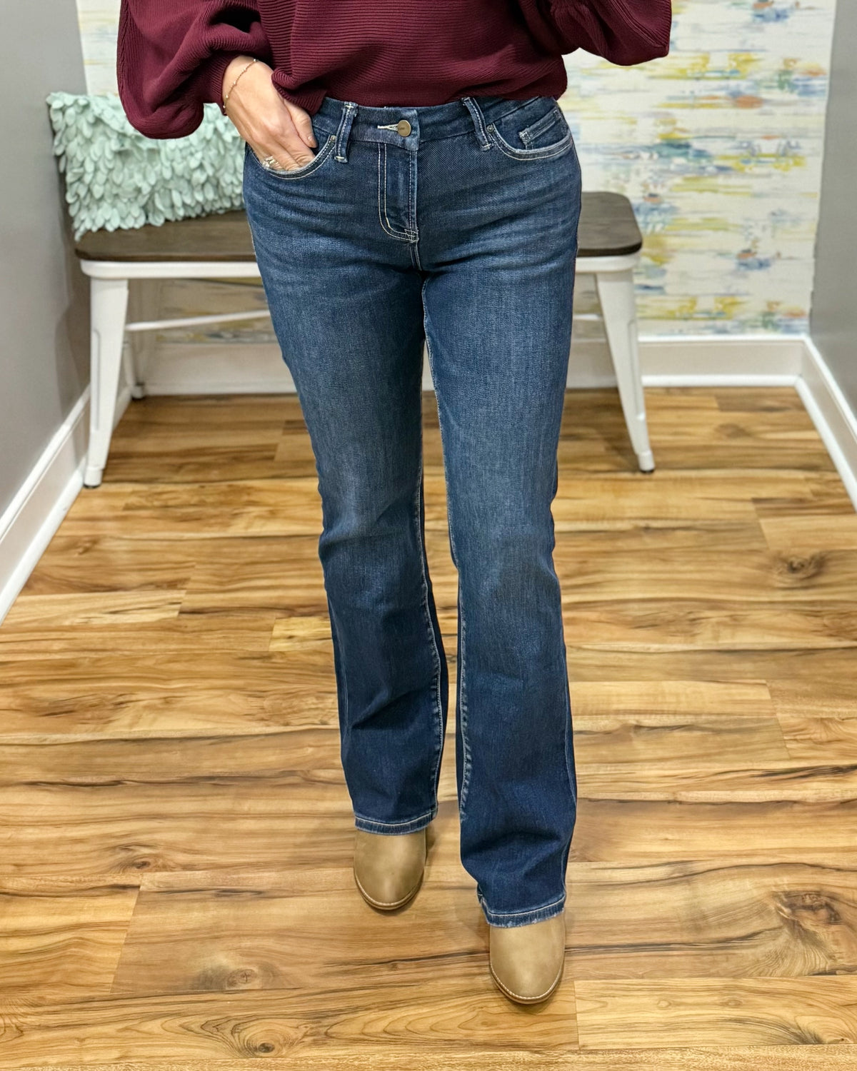 A person wearing mica dark blue bootcut jeans, a burgundy top, and tan boots, standing on a wood floor in front of a white bench and a colorful wall.