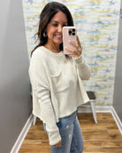 A woman takes a mirror selfie wearing a cream-colored, textured knit long-sleeve crop top with a chest pocket, paired with blue jeans.