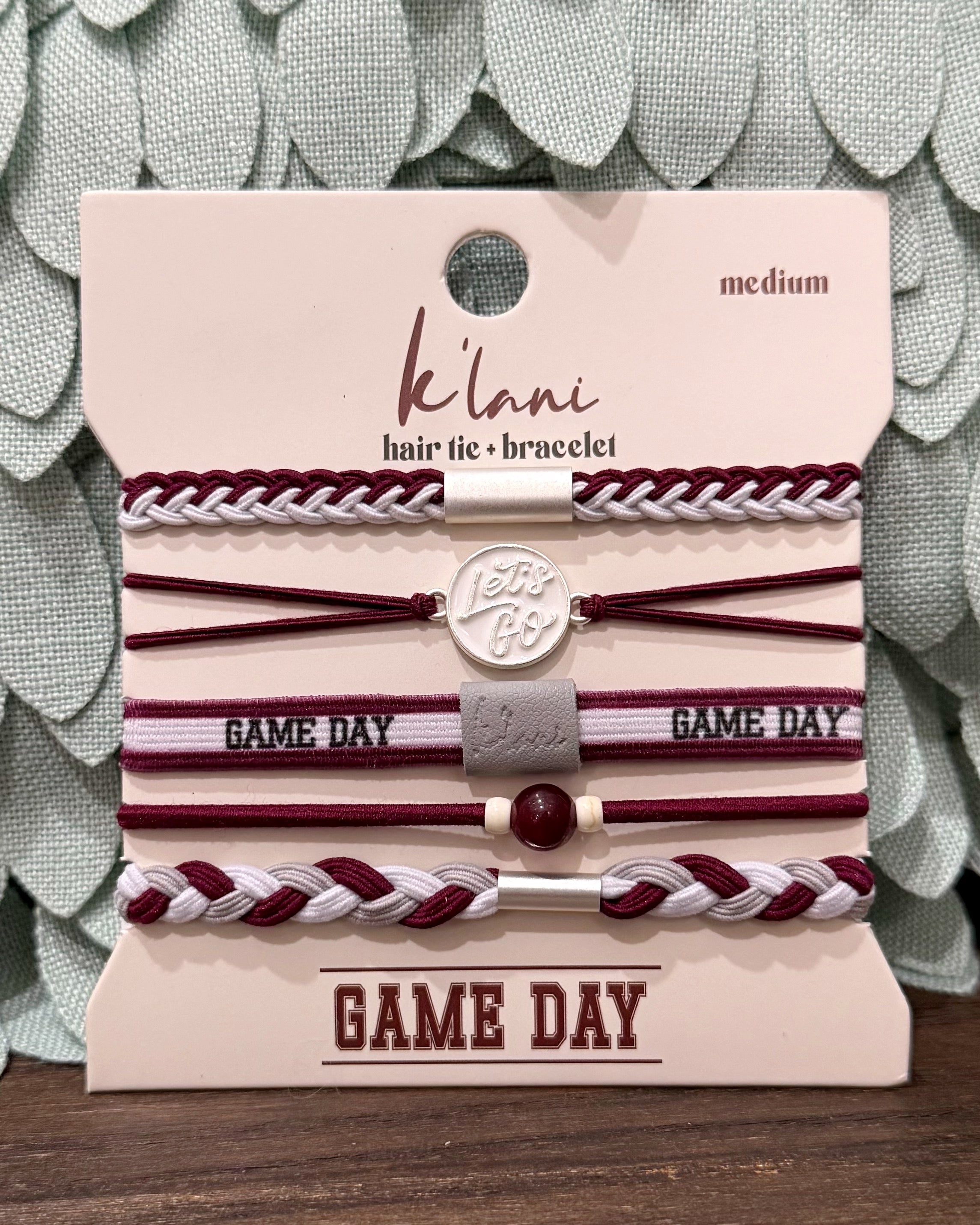 A set of six K'lan hair tie and bracelet accessories on a branded display card, featuring burgundy, white, and silver elements with 'GAME DAY' text, photographed against a textured green background.