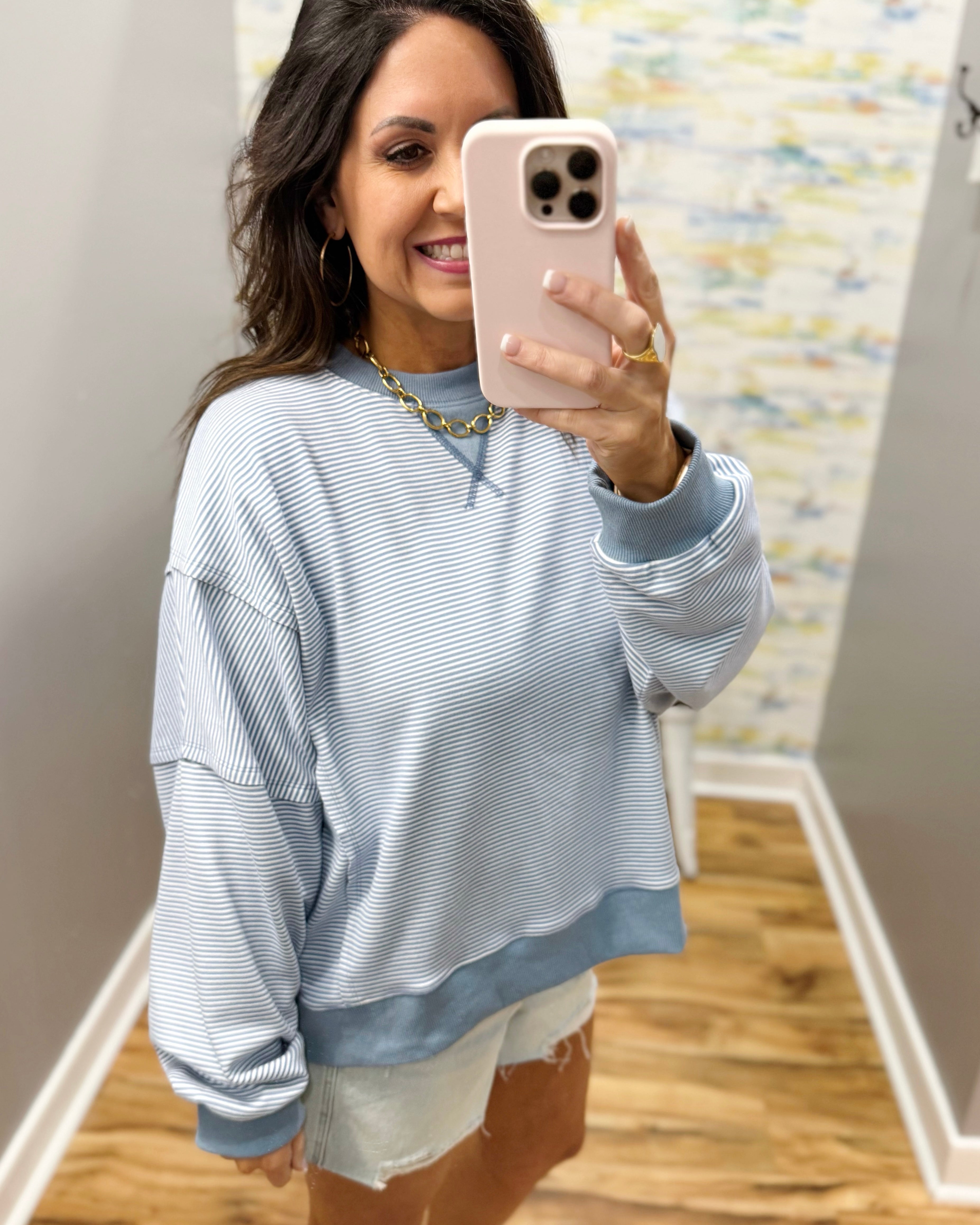 Woman taking a mirror selfie wearing a light blue striped sweater and white shorts.