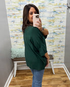 A woman takes a mirror selfie wearing an oversized dark green short-sleeve top and blue jeans in a room with a patterned wallpaper background and a wooden bench.