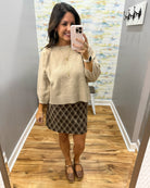 A woman takes a mirror selfie in a fitting room wearing a beige knit crewneck sweater, a brown and black plaid mini skirt, and Corkys Pity Party shoes in Cognac.