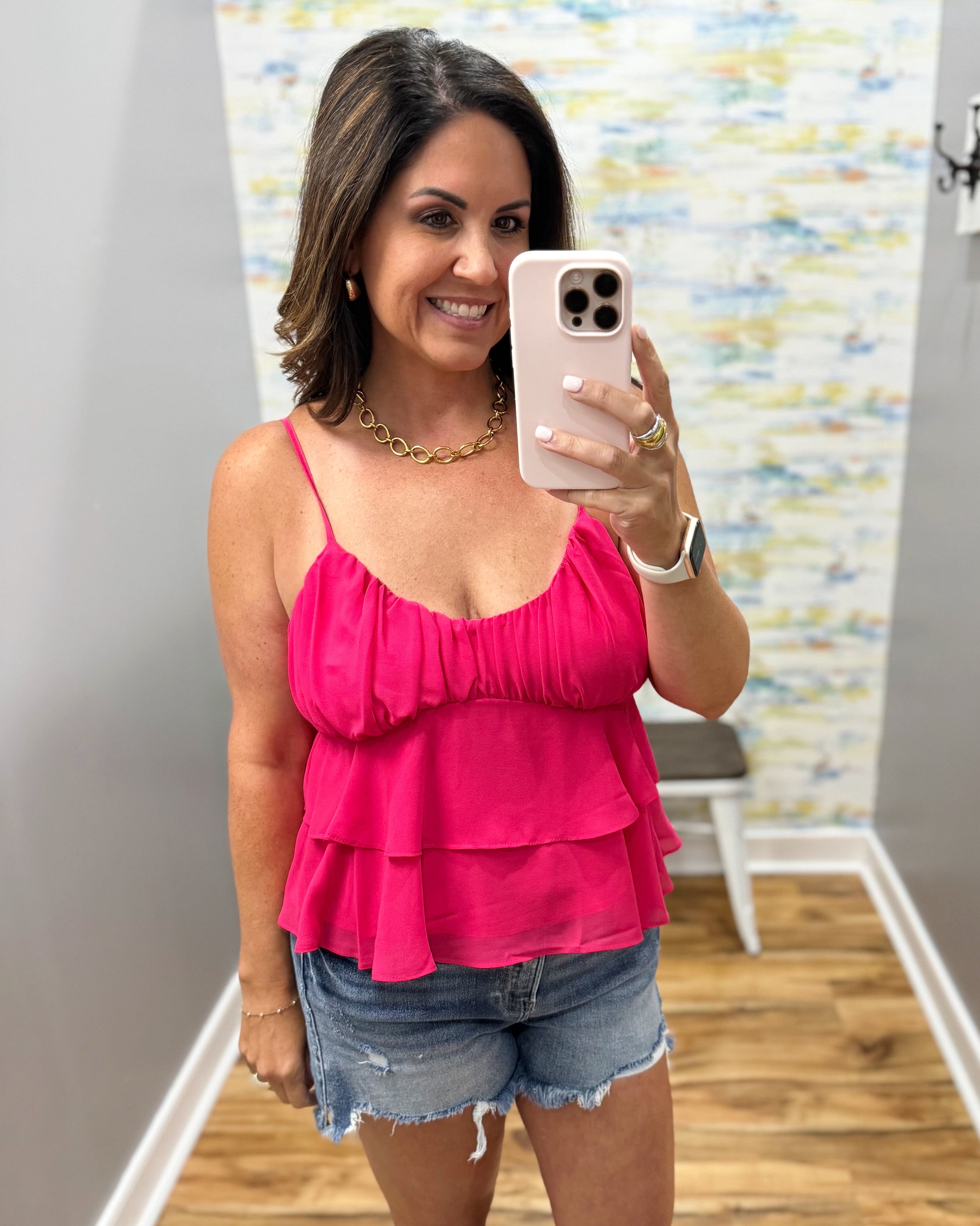 Woman taking a mirror selfie wearing a pink top and denim shorts in a room with a colorful wall.