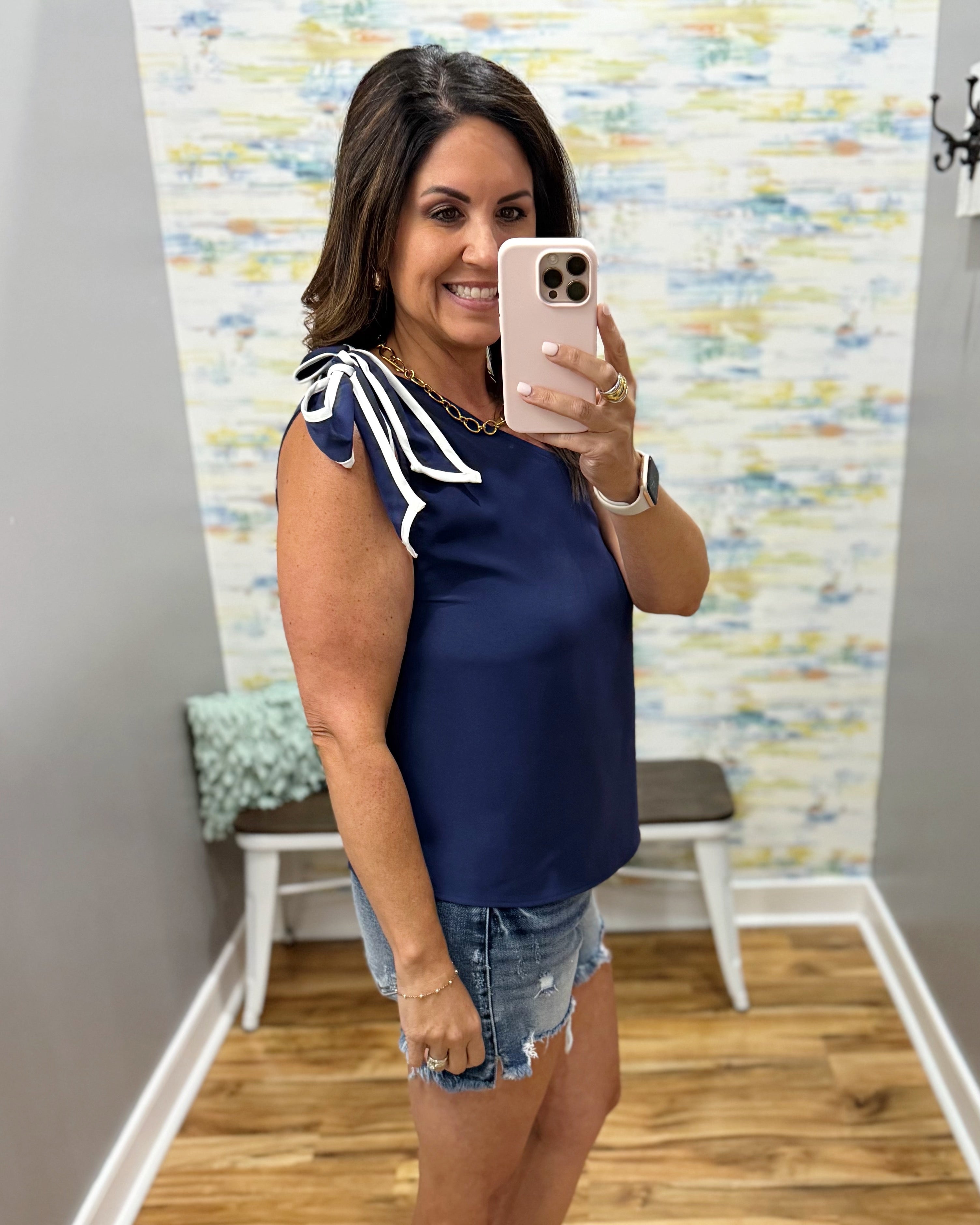 Woman taking a mirror selfie wearing a navy blue top with white accents and denim shorts.