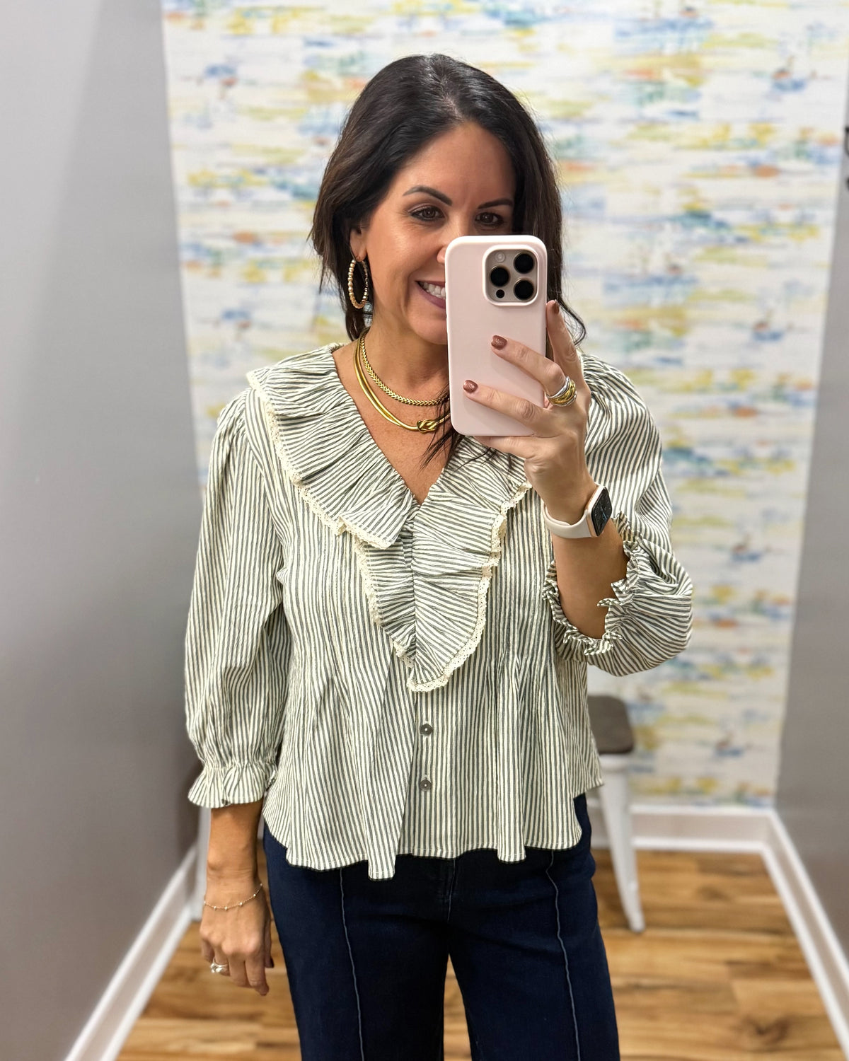 A woman takes a mirror selfie wearing a light sage green and white vertically striped blouse with a ruffled collar and front buttons, paired with dark jeans.