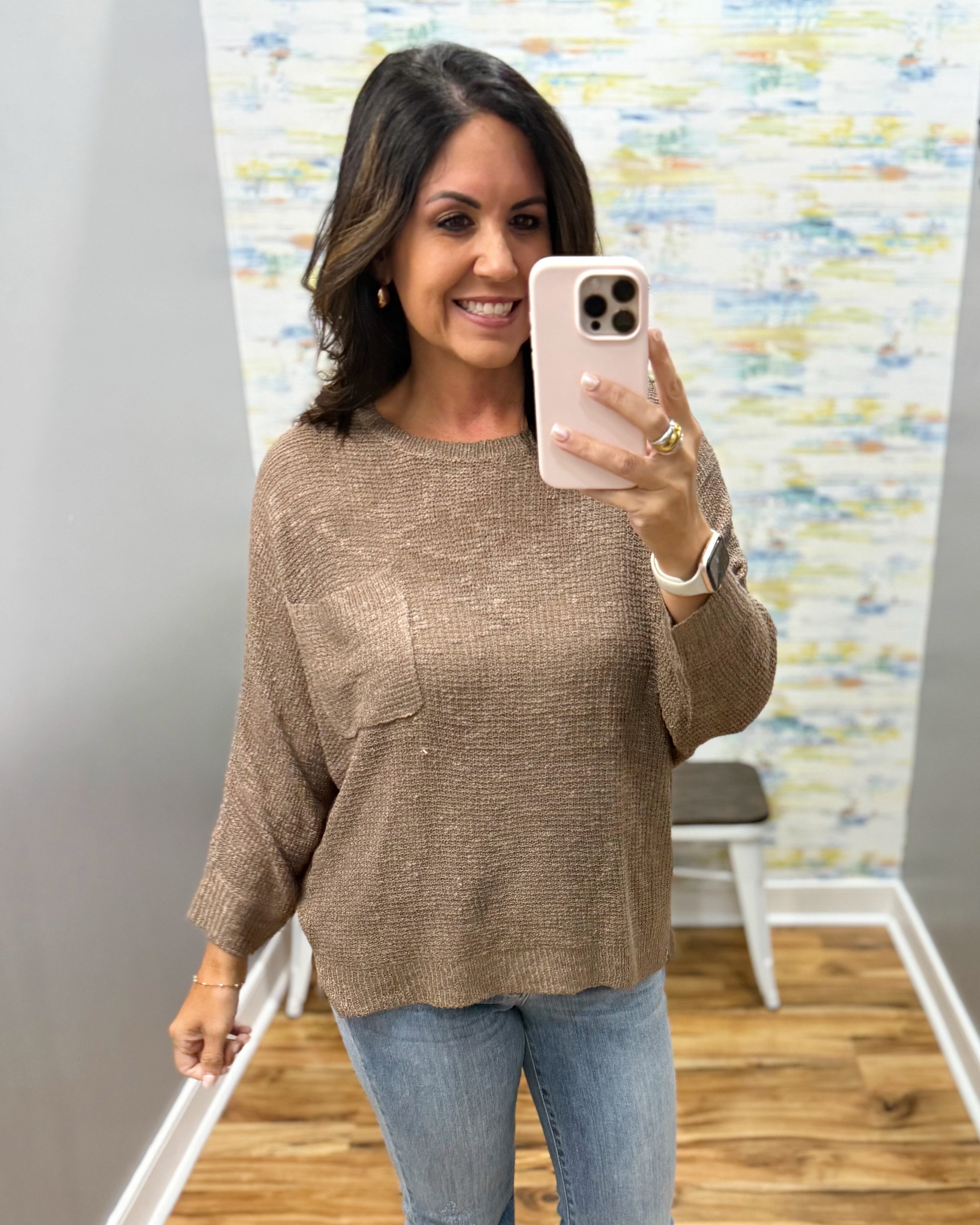 A woman takes a mirror selfie wearing a textured, waffle-knit long-sleeve top in a taupe brown color, paired with blue jeans. The setting appears to be a boutique fitting room with a wooden floor and abstract wall art.