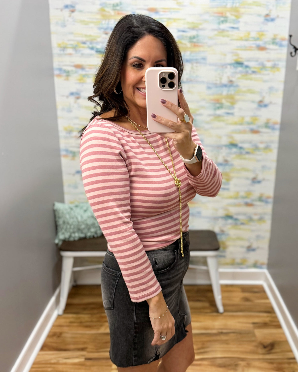 A woman taking a mirror selfie in a fitting room wearing a ginger and mauve striped long-sleeve top and a dark gray denim skirt.