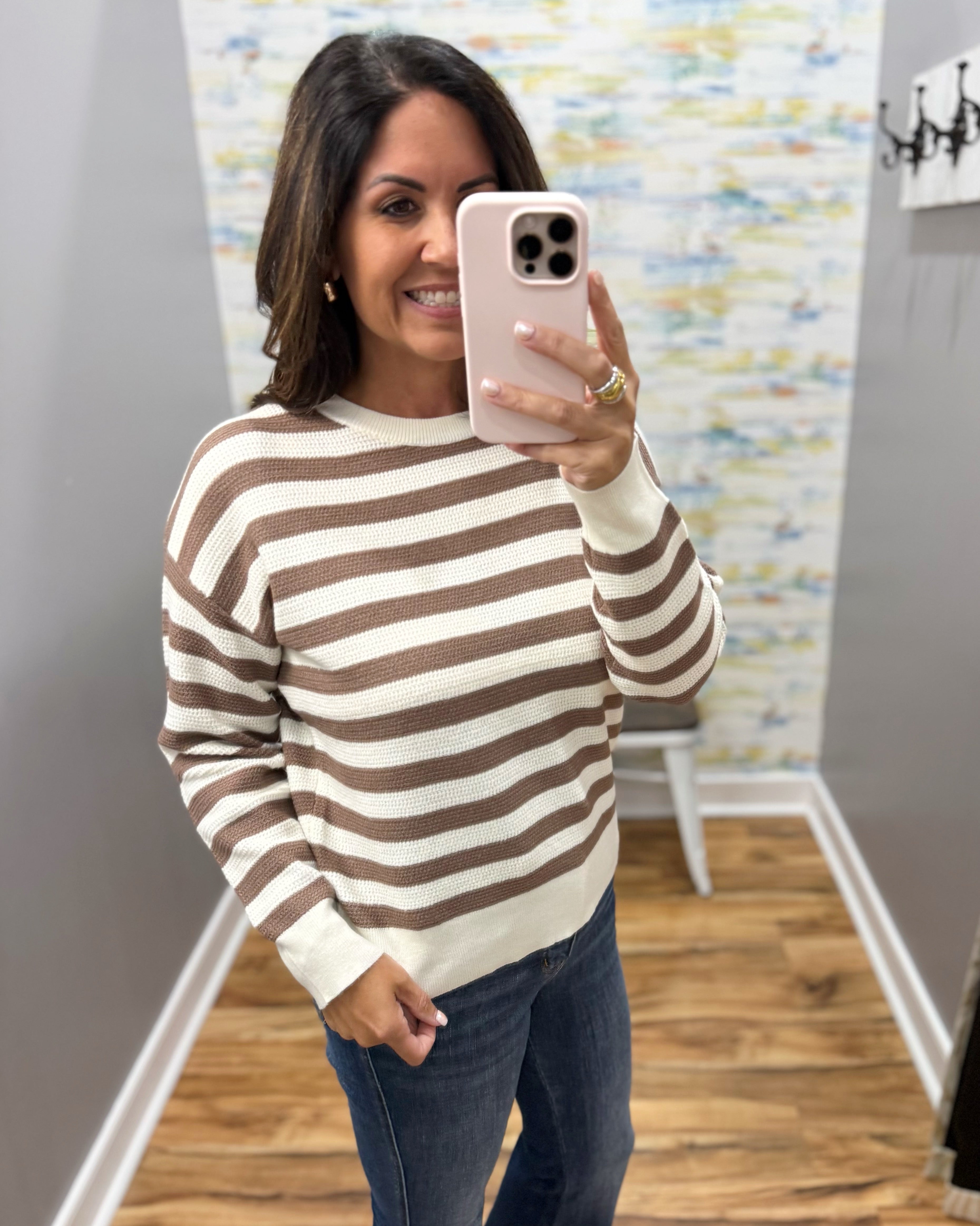 A woman takes a mirror selfie wearing a cream and brown horizontally striped long-sleeve knit sweater and dark blue jeans in a home hallway.