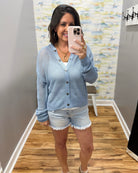Woman taking a mirror selfie wearing a light blue cardigan and denim shorts in a room with patterned wallpaper.