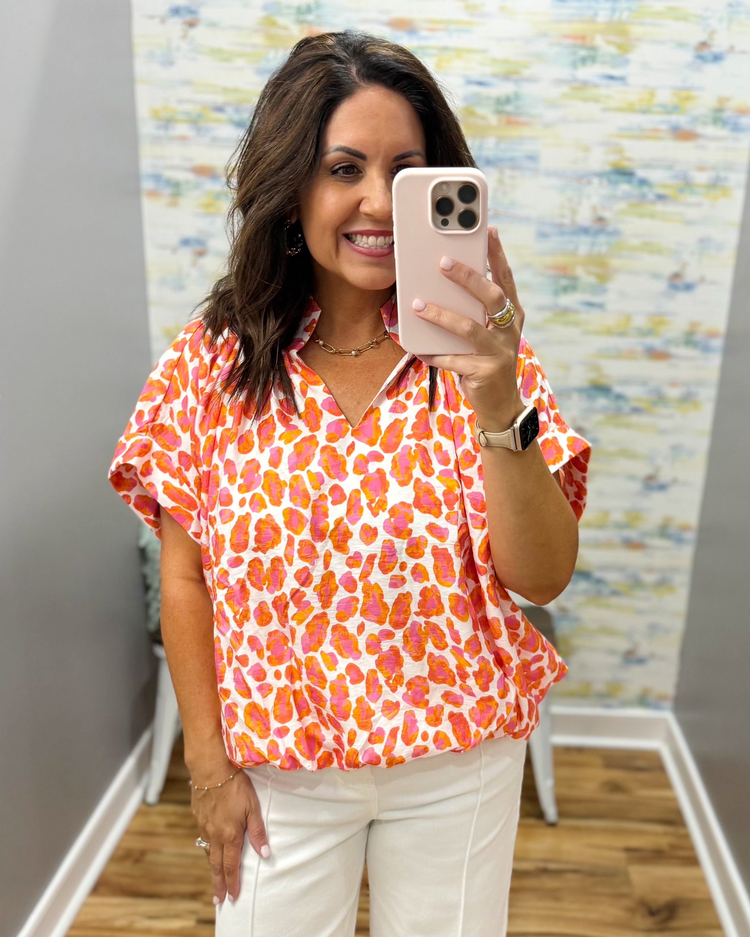 Woman taking a mirror selfie wearing an orange and pink patterned blouse and white pants.