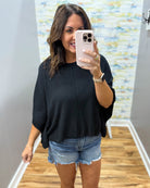 Woman taking a mirror selfie wearing a black top and denim shorts in a room with a colorful wall.