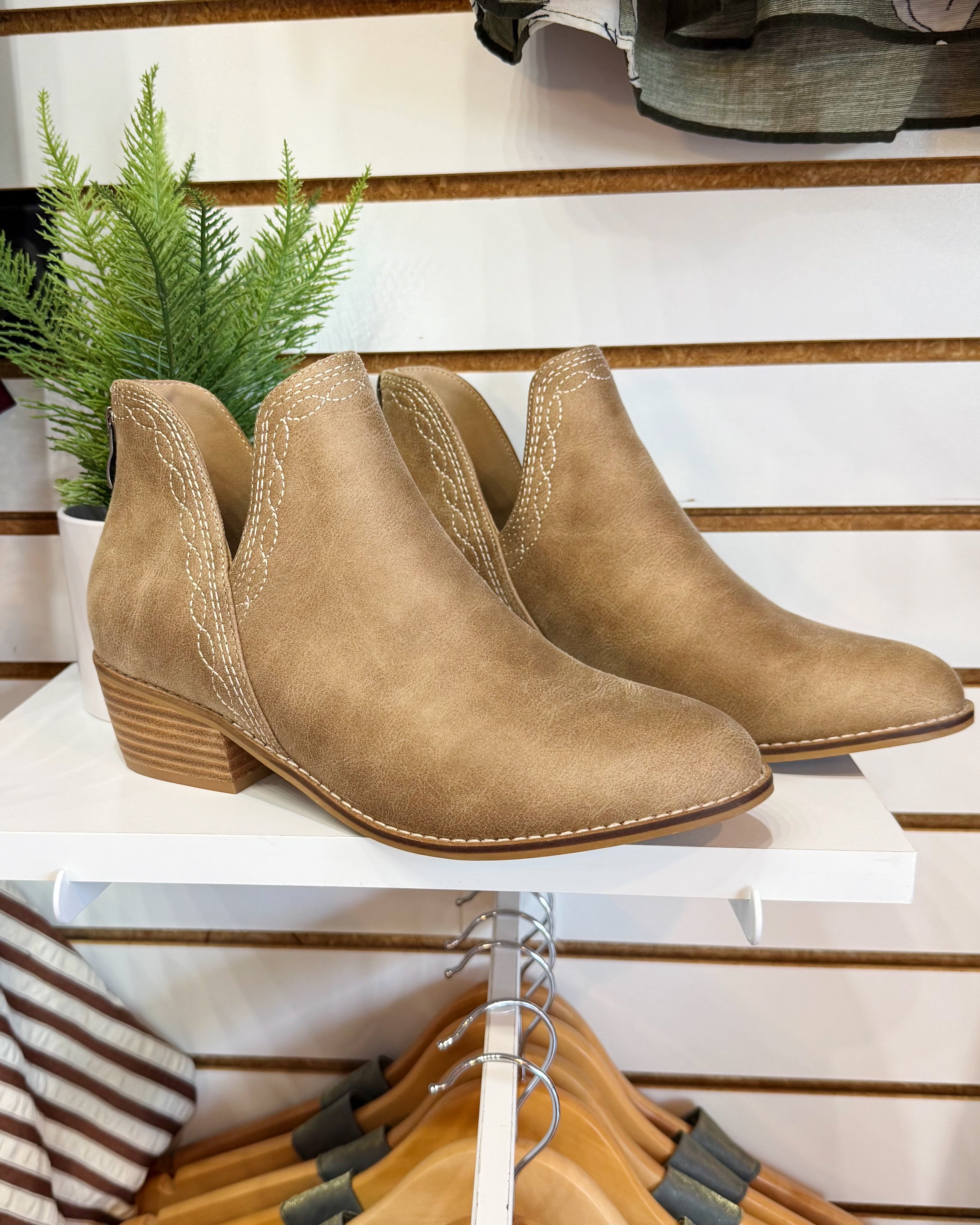 A pair of tan suede ankle boots with stacked block heels and contrast stitching, displayed on a white shelf in a retail setting.