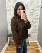 A woman takes a mirror selfie wearing a brown textured knit sweater and blue jeans in a home setting with a patterned wallpaper background.