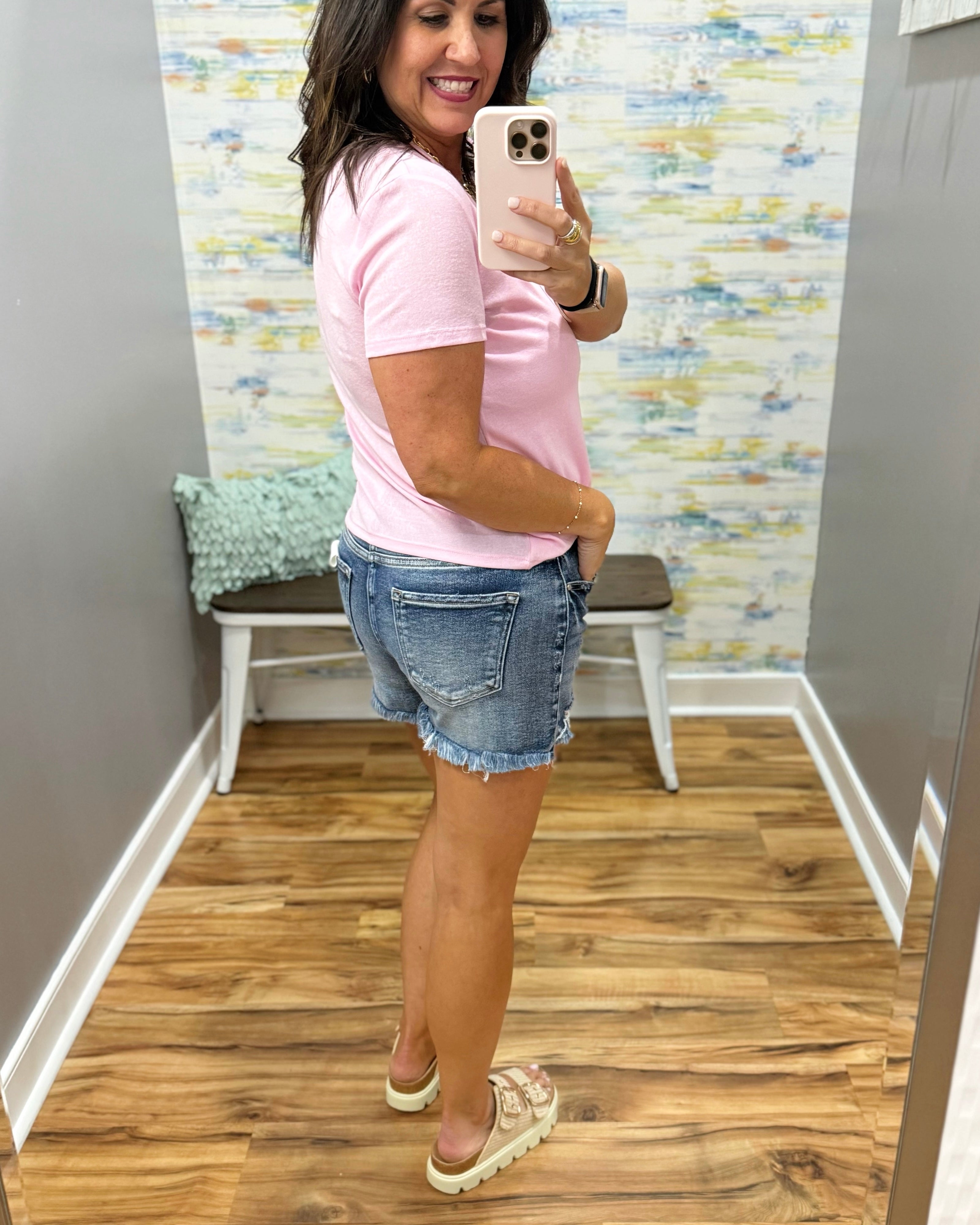 Woman taking a mirror selfie wearing a pink t-shirt and denim shorts in a room with a colorful wall.