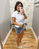 Woman taking a mirror selfie wearing a white striped shirt and denim shorts in a room with patterned wallpaper.