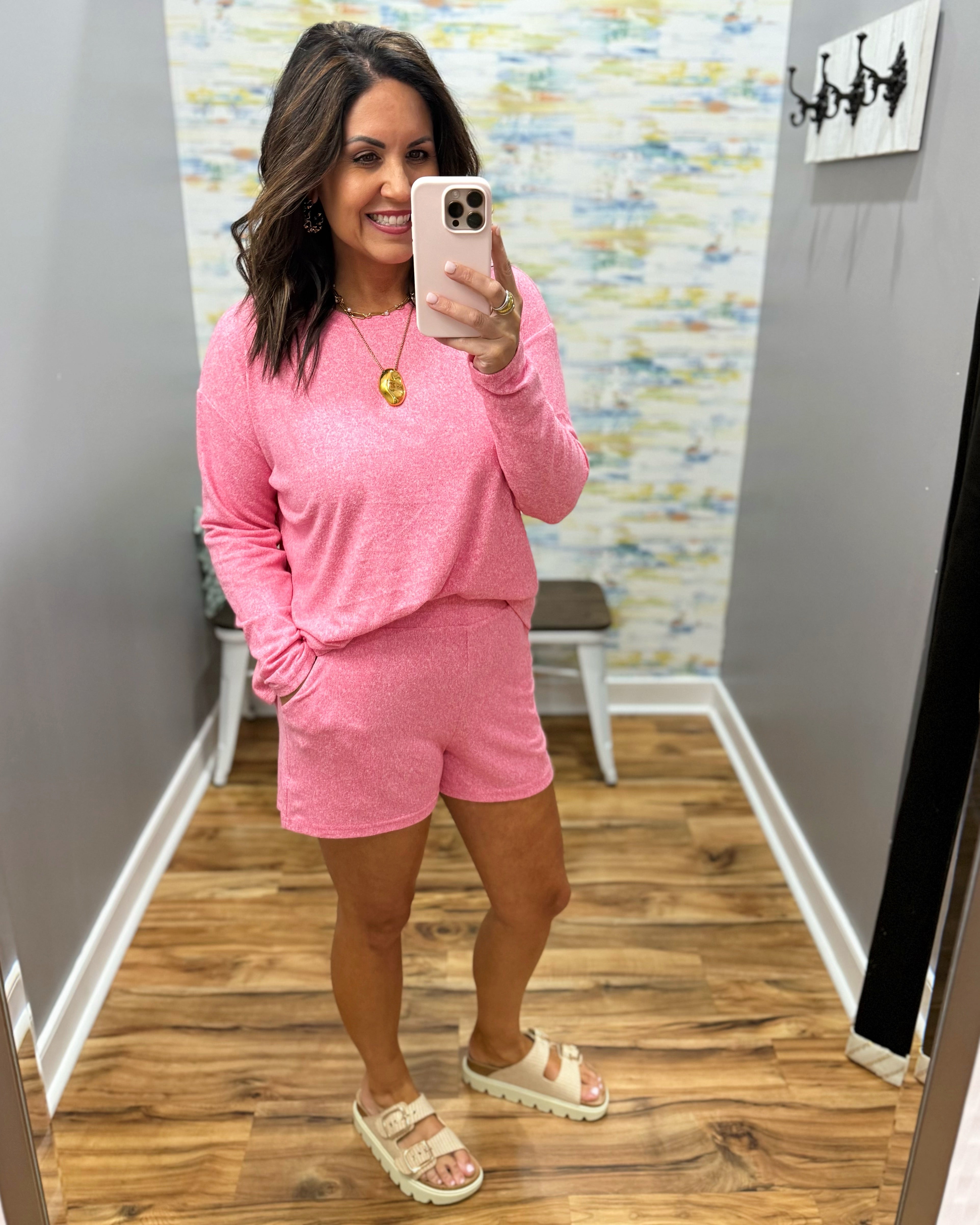 Woman taking a mirror selfie wearing a pink outfit in a room with a patterned wall.