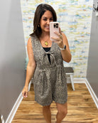 A woman takes a mirror selfie wearing a sleeveless leopard-print romper with a tie detail at the chest over a black top. The setting is a home fitting room with wood floors, gray walls, and a colorful abstract wallpaper.